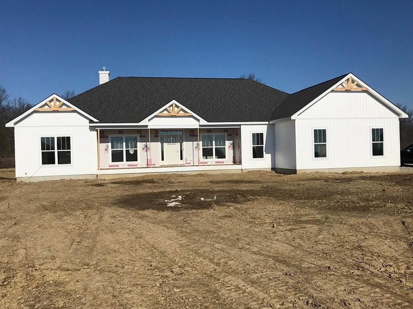 Modern house under construction with black roof, white siding, glass-paneled door, large windows, and dirt field with tire tracks in foreground