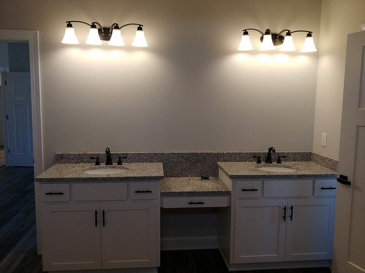 Bathroom with dual sinks set in a marble countertop, white cabinetry with black handles, wall-mounted light fixtures above mirrors, and a white door with a black handle.
