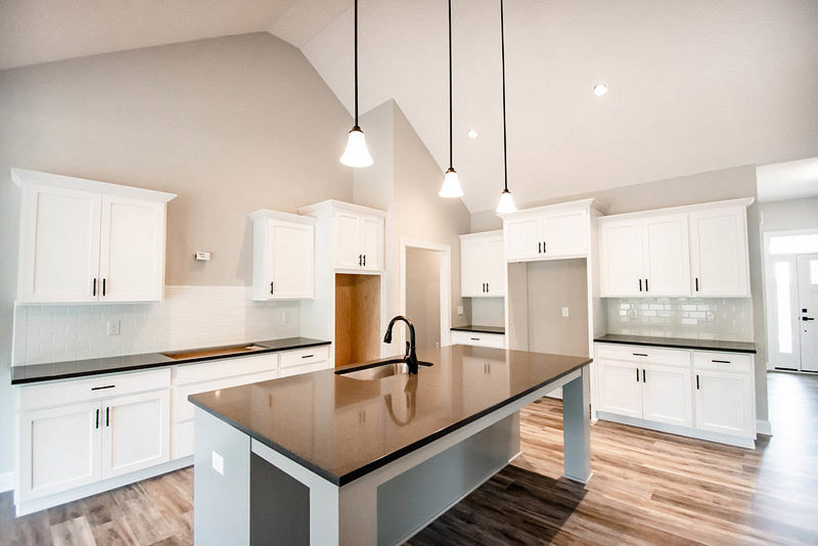 Kitchen with black countertop, central island featuring built-in sink and faucet, white cabinetry, tiled backsplash, bell-shaped pendant light, stainless steel appliances, light