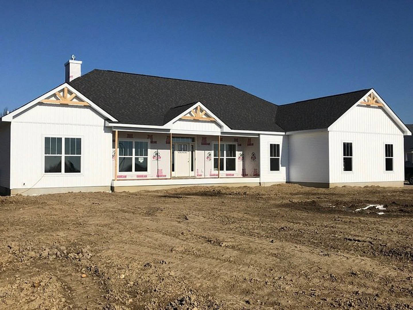 Two-story house under construction with exposed framing, dirt yard, white siding, white door featuring a red logo, and blue sky in the background