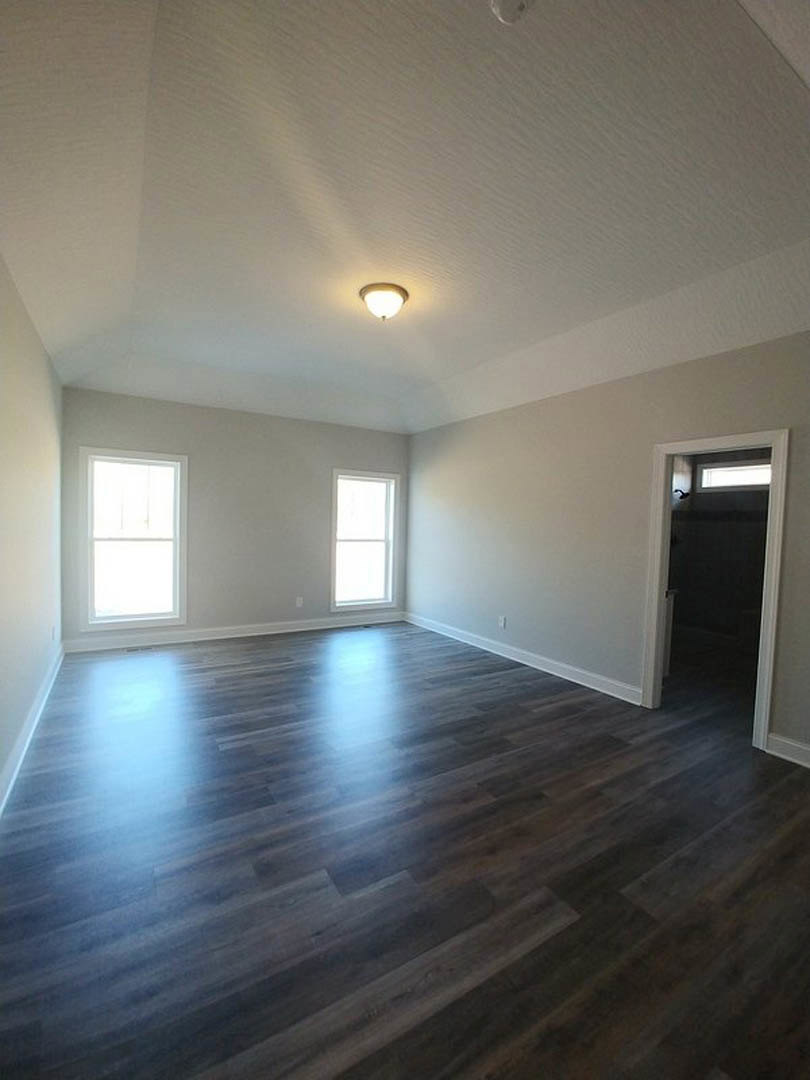 Wood flooring in a bright room with two windows, plaster walls, ceiling-mounted light fixture, and a door leading to a darker space