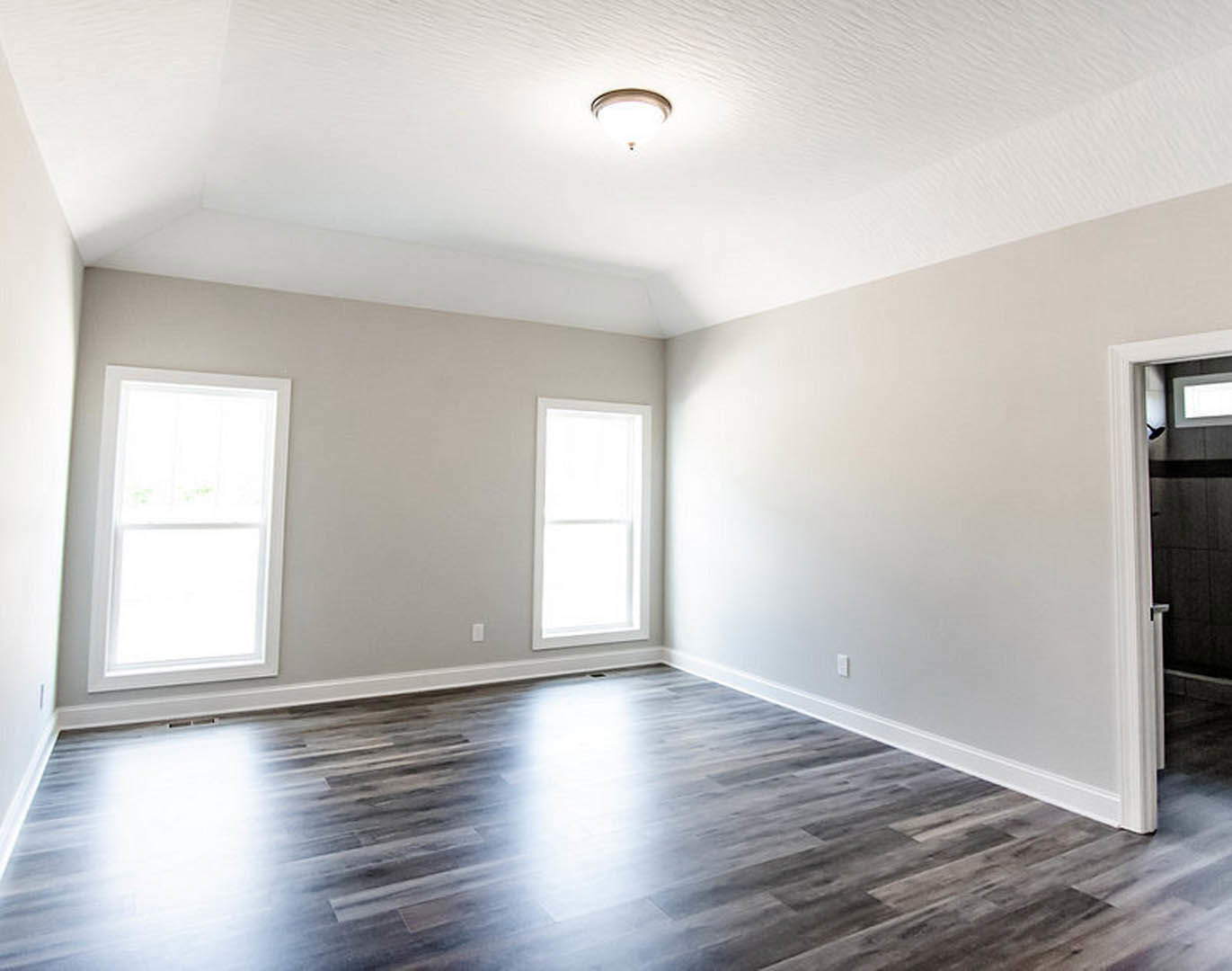 Spacious room with warm wood flooring, two large windows, white walls, and ceiling light fixture illuminating the floor.