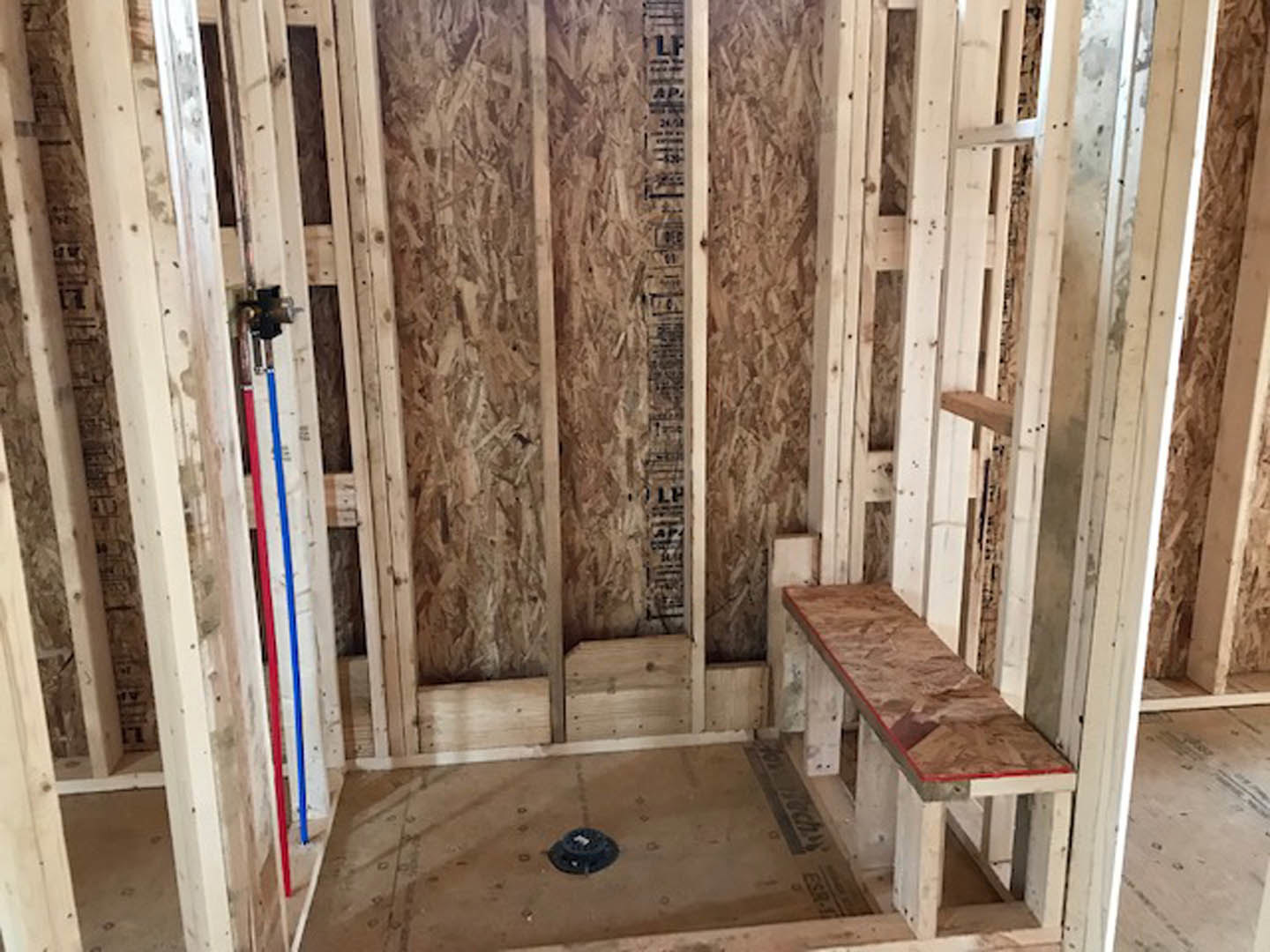 Unfinished bathroom with exposed wood framing, plywood walls, wooden bench, black circular drain on brown floor, and partial brick wall visible