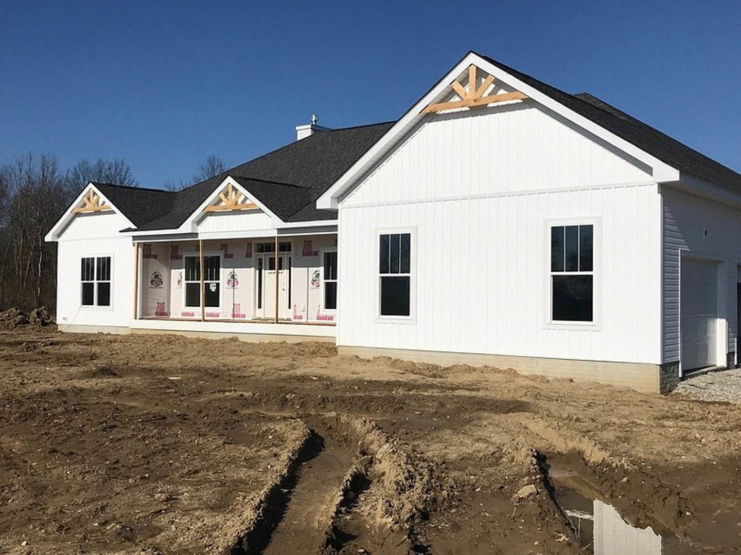 White siding house under construction with visible window frames, dirt ground marked by tire tracks, and clear blue sky overhead