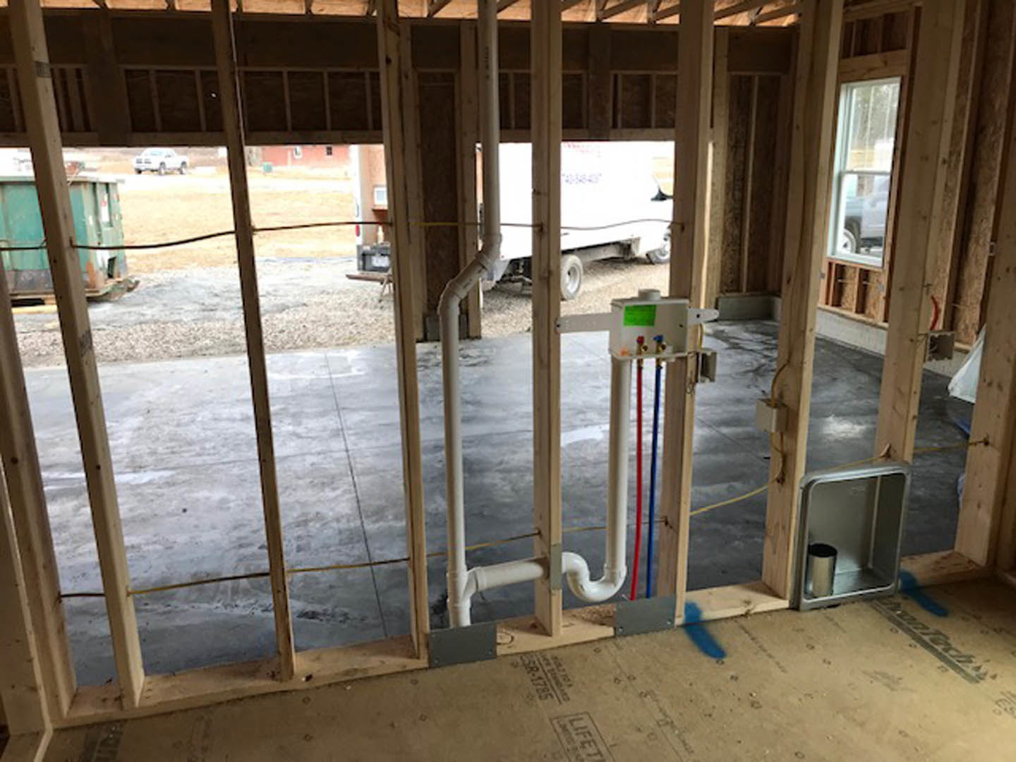 Unfinished room with exposed wooden beams, white pipes along a wall, large window, and construction materials scattered on the floor