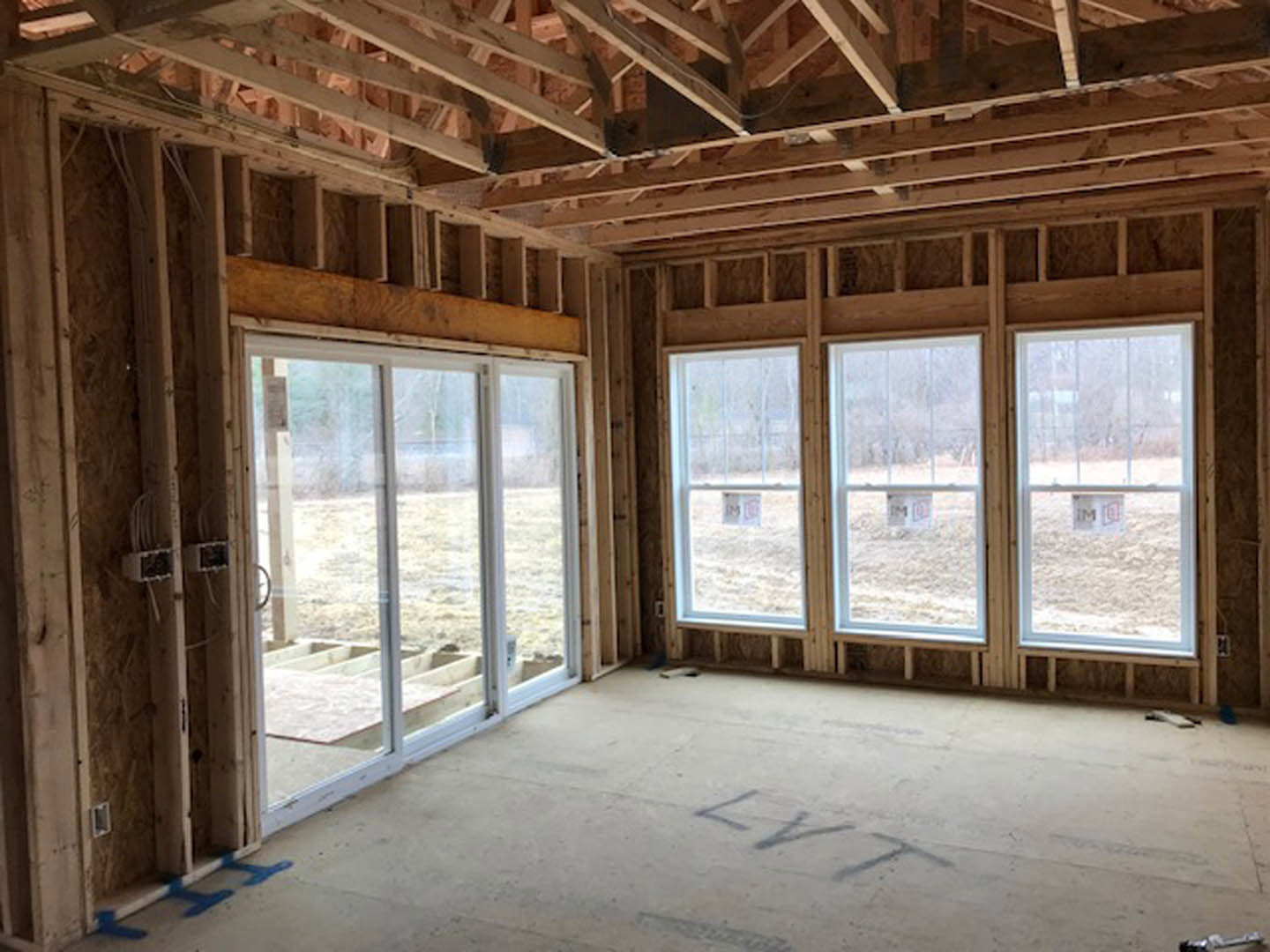 Spacious room with large windows, exposed wood beams, concrete floor with blue paint accents, glass door, and sunlight casting a person’s shadow across the ground