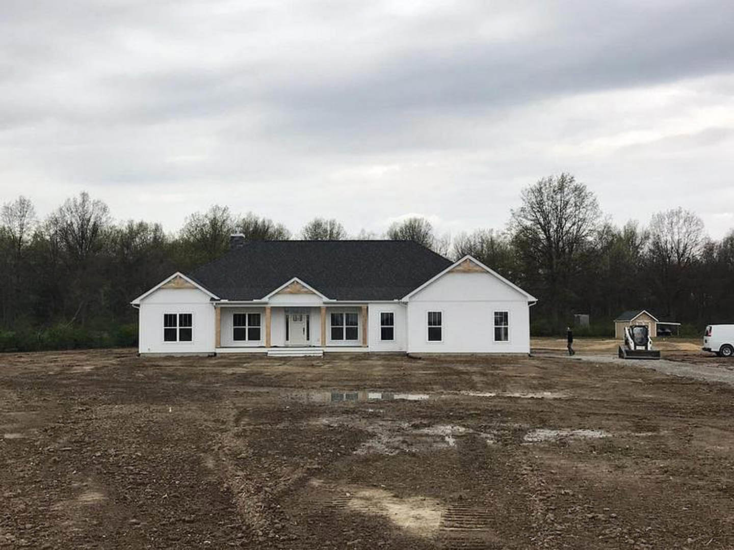 White house with black roof under construction, surrounded by dirt field, white van parked on nearby road, group of trees in background, partially visible bulldozer, cloudy sky