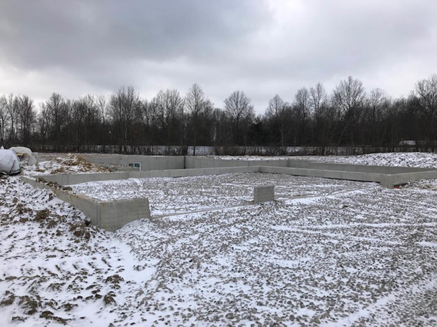 Concrete foundation slab surrounded by snow and scattered rocks in an open field, leafless trees and cloudy winter sky in the background