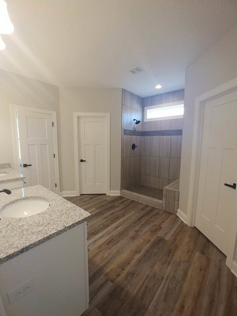 Bathroom featuring two white sinks with stone countertops, glass-enclosed tile shower, white cabinetry, black hardware, and wall-mounted light fixtures.