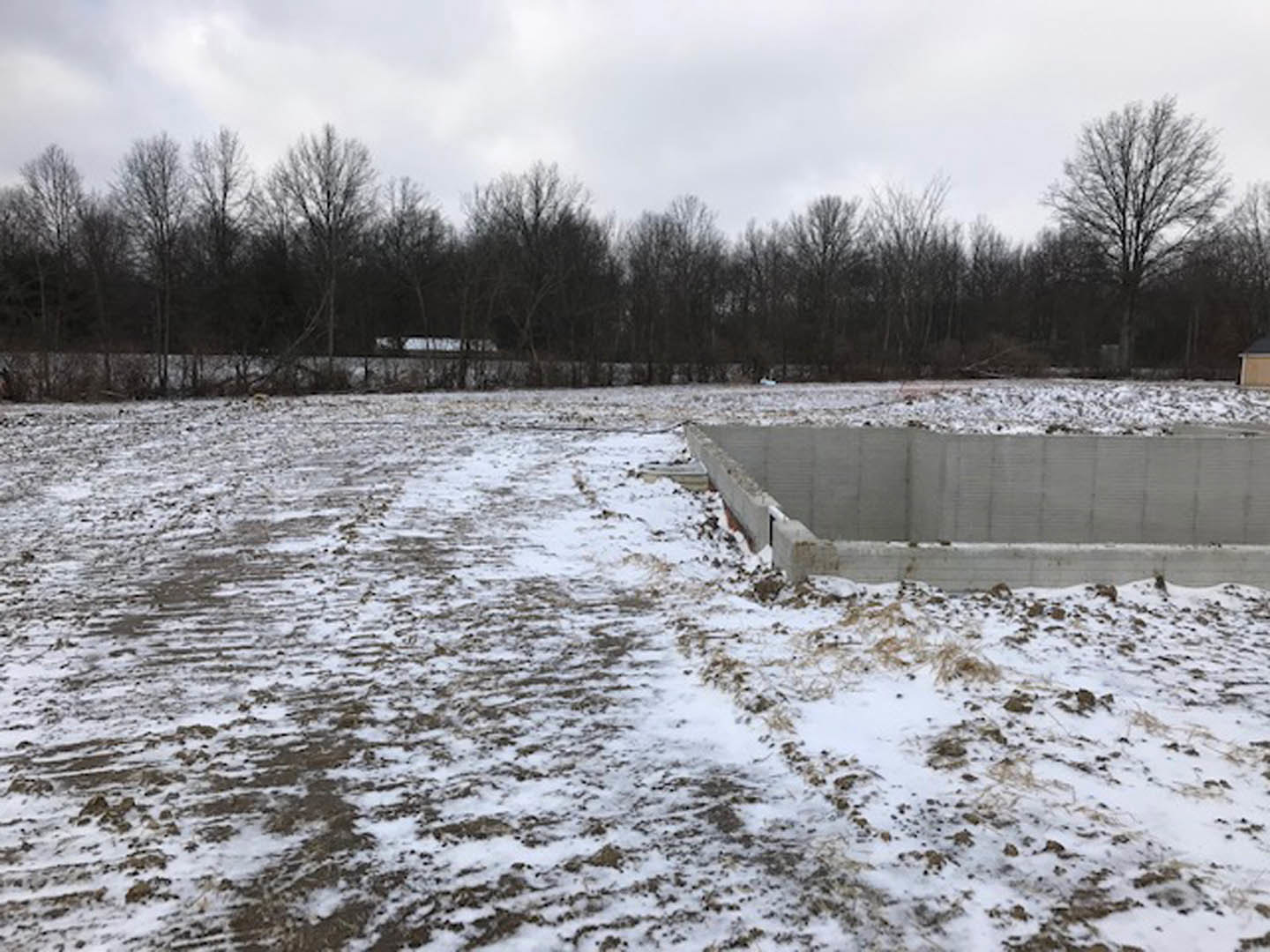 Concrete foundation set in a snow-covered field, leafless trees in the background, footprints visible on the snowy ground