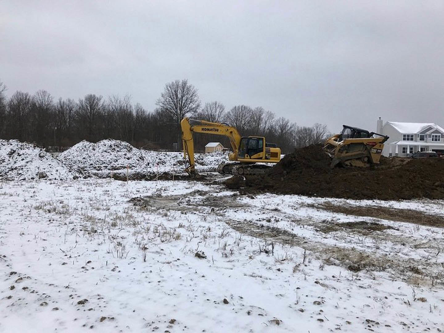 Yellow bulldozer parked beside a mound of dirt on a snowy construction site, bare trees in background, snow covering ground and nearby unfinished house