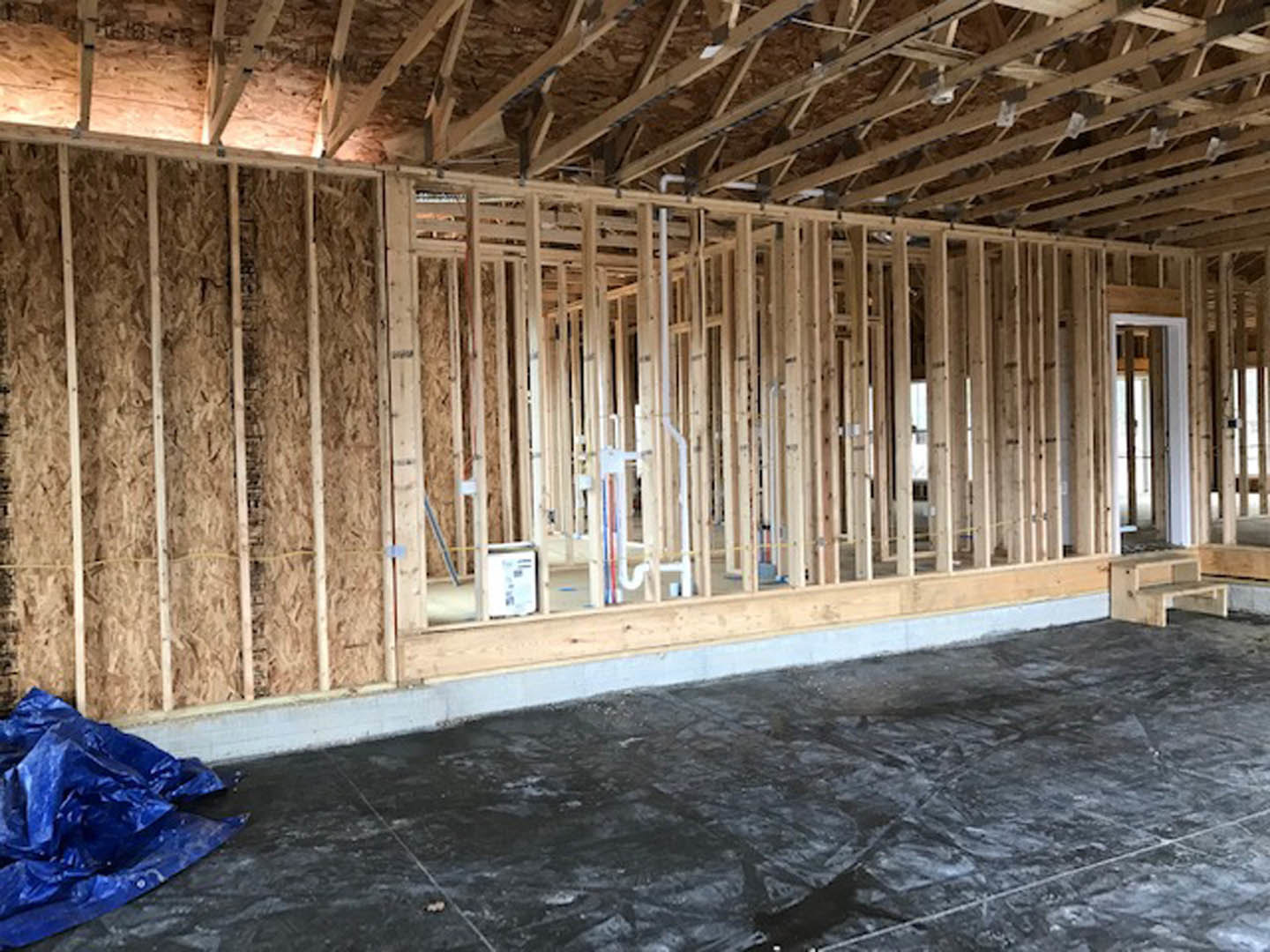 Wood-framed room under construction with exposed beams, chipboard wall, black subfloor, and blue tarp on the ground