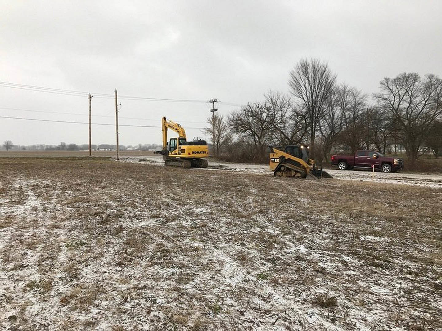 Yellow excavator and white pickup truck parked on snowy field with bare trees and cloudy sky in background