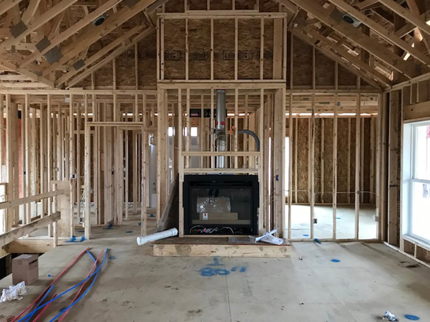 Living room with exposed wood framing, built-in fireplace with glass window, white-framed window, red and blue electrical wires on unfinished floor, silver hardware, and white