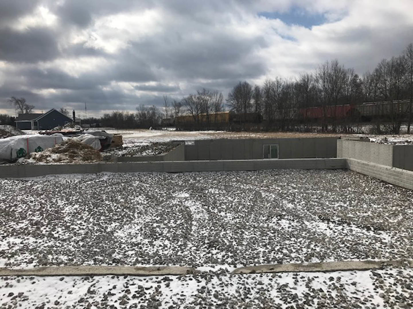 Two-story home under construction with white-framed windows, exposed wooden framing, and snow covering the ground and roof, surrounded by bare trees.