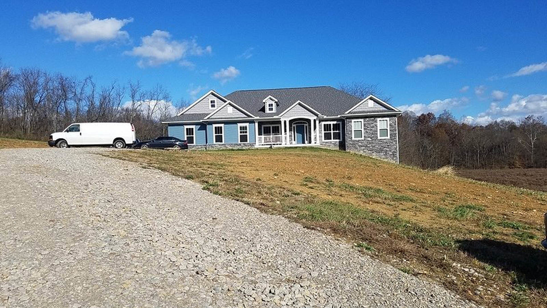 Modern cottage-style home with light siding and gabled roof, situated on a grassy hill with a white van parked in front, surrounded by trees and open sky.