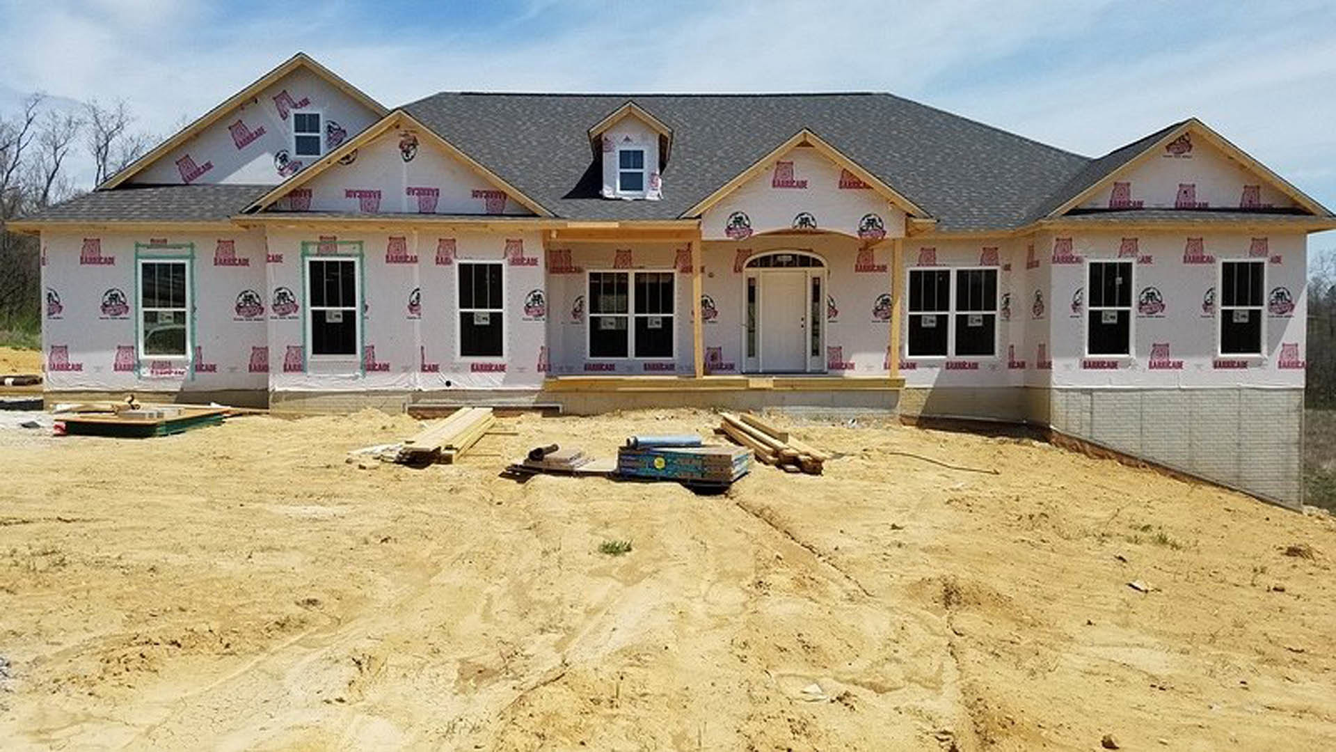 Partially built house with exposed framing, dirt yard, white door with glass panels, multiple white-framed windows including one with a sign, cloudy sky overhead, blurred car
