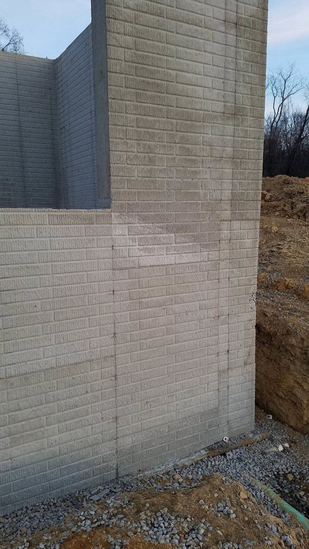 Red brick exterior wall with a rectangular opening, surrounded by stone accents, tree branches visible against blue sky above, pile of rocks on ground below