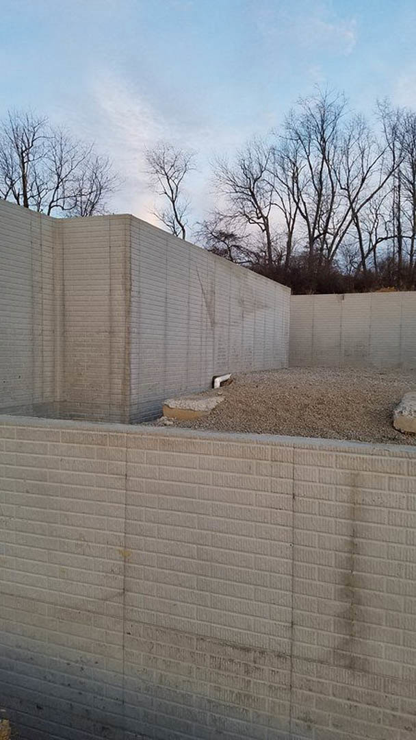 Concrete retaining wall bordered by gray gravel, exposed pipe along base, leafless trees and blue sky in background