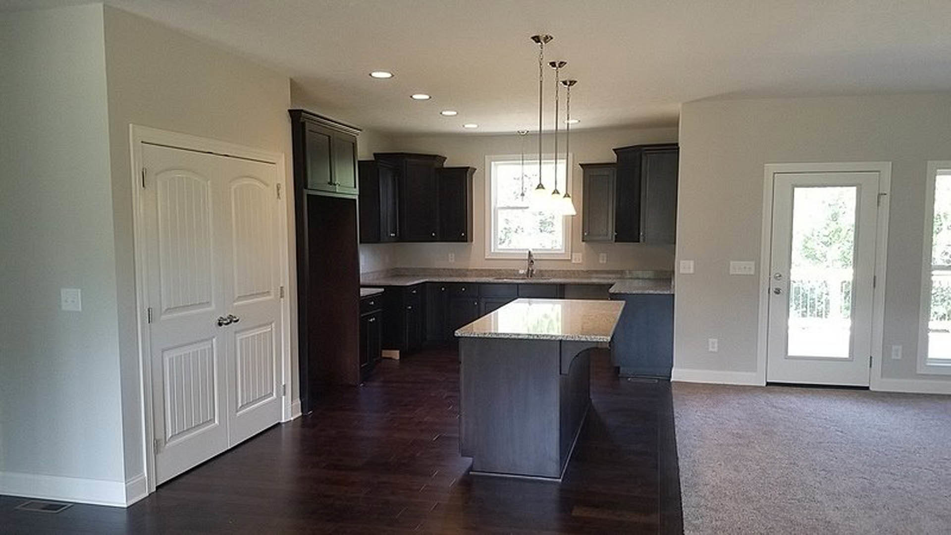 Kitchen with dark wood cabinets, marble-topped island, laminate flooring, white wall switch, white door with glass panel, and carpeted adjacent room