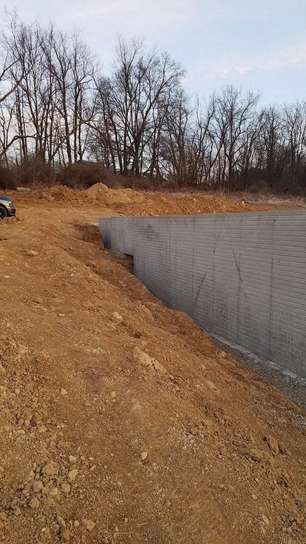 Concrete retaining wall set into a dirt hillside, surrounded by leafless trees and sparse grass, with a large hole excavated in the foreground