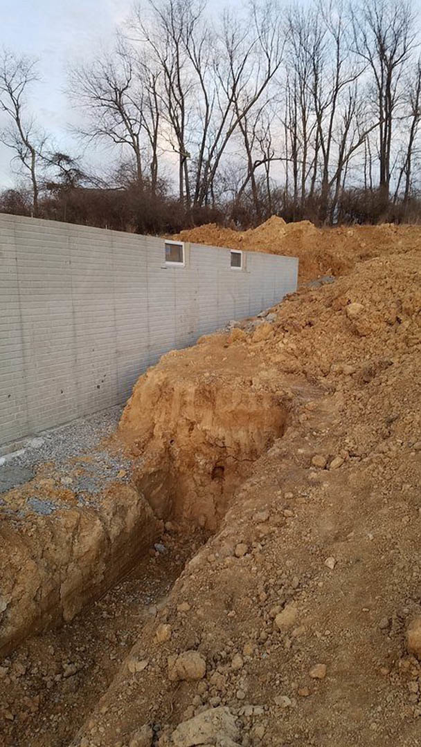 Excavated foundation surrounded by piles of soil, bare trees in the background, exposed rock wall along the edge