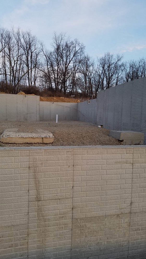 Smooth concrete wall with visible foundation, wire fencing along the perimeter, trees and cloudy sky in the background