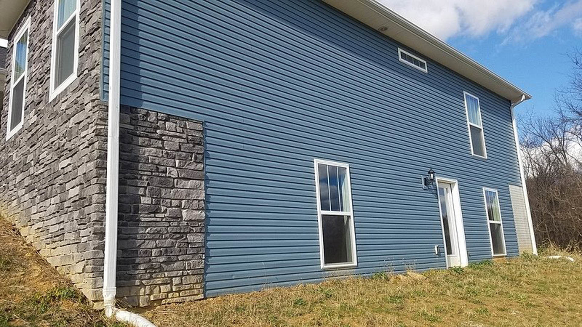 Blue siding exterior with white-framed window and stone accent wall, surrounded by grass under a partly cloudy sky.