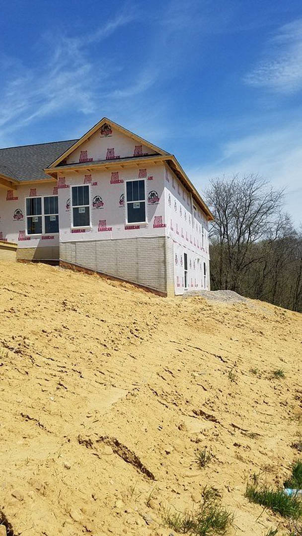 Partially built house with exposed framing and windows, situated on a steep dirt slope, surrounded by trees and cloudy sky