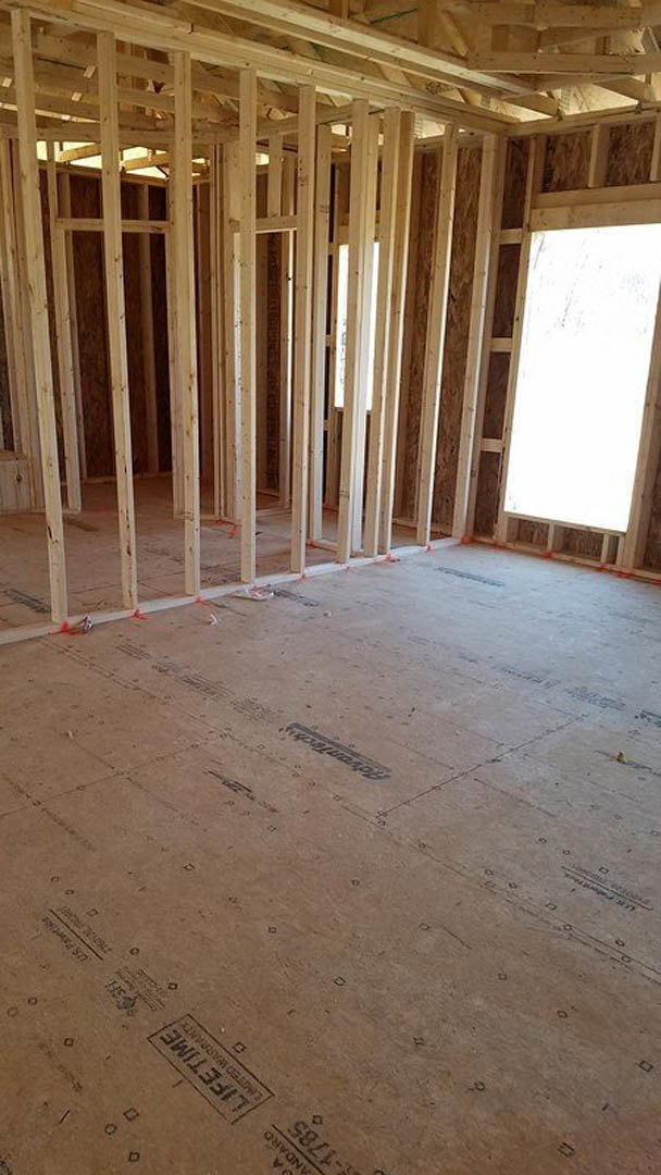 Room under construction with exposed wood framing, unfinished walls, window opening with white background, visible ceiling beams, and construction materials on the floor.