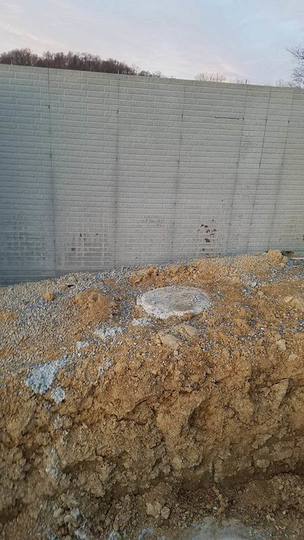 Grey brick retaining wall bordered by dirt and gravel, with scattered rocks and distant trees visible under a cloudy sky.