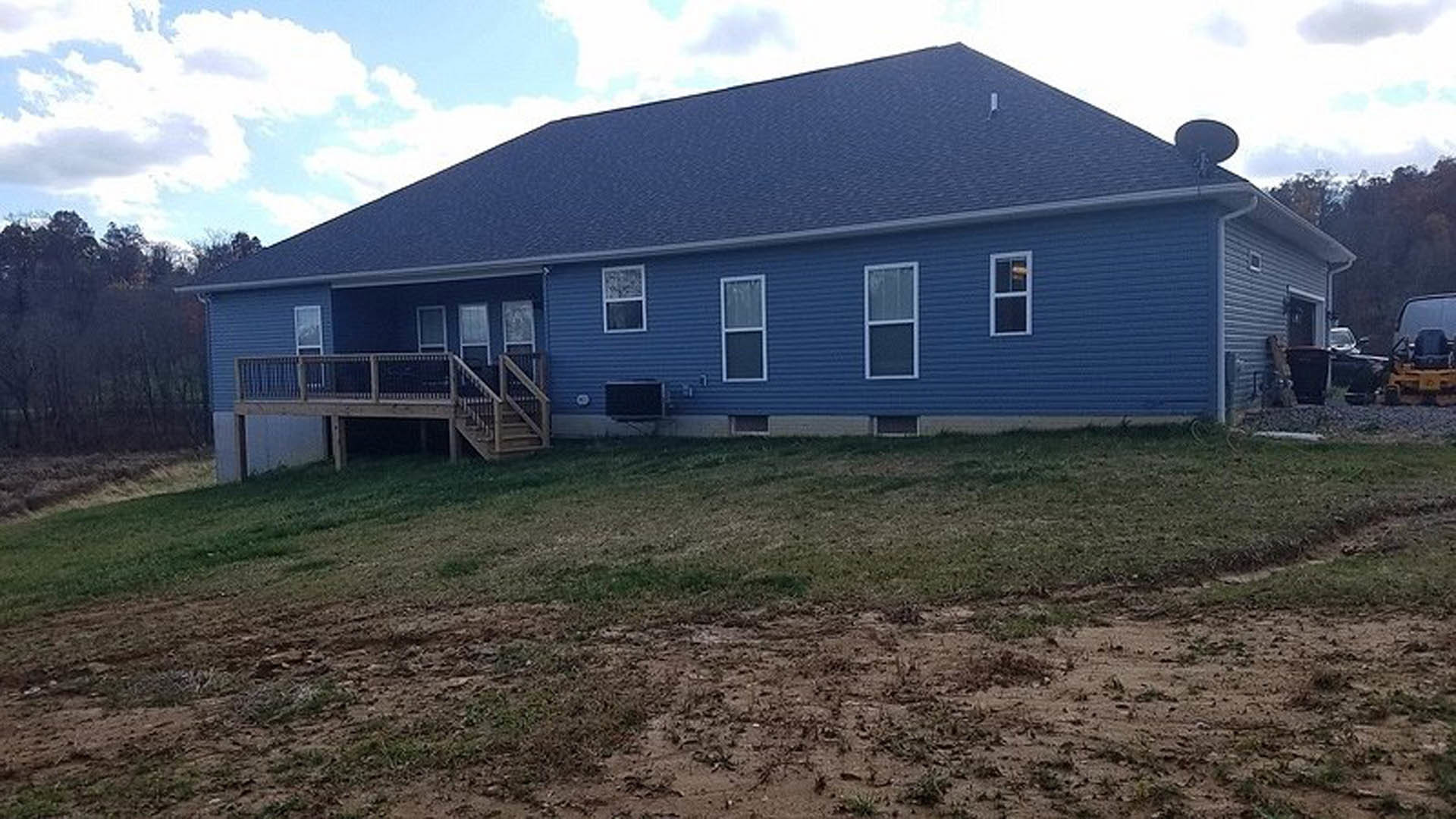 Blue cottage-style home with white trim, elevated wooden deck, grassy yard, and scattered trees under a cloudy sky
