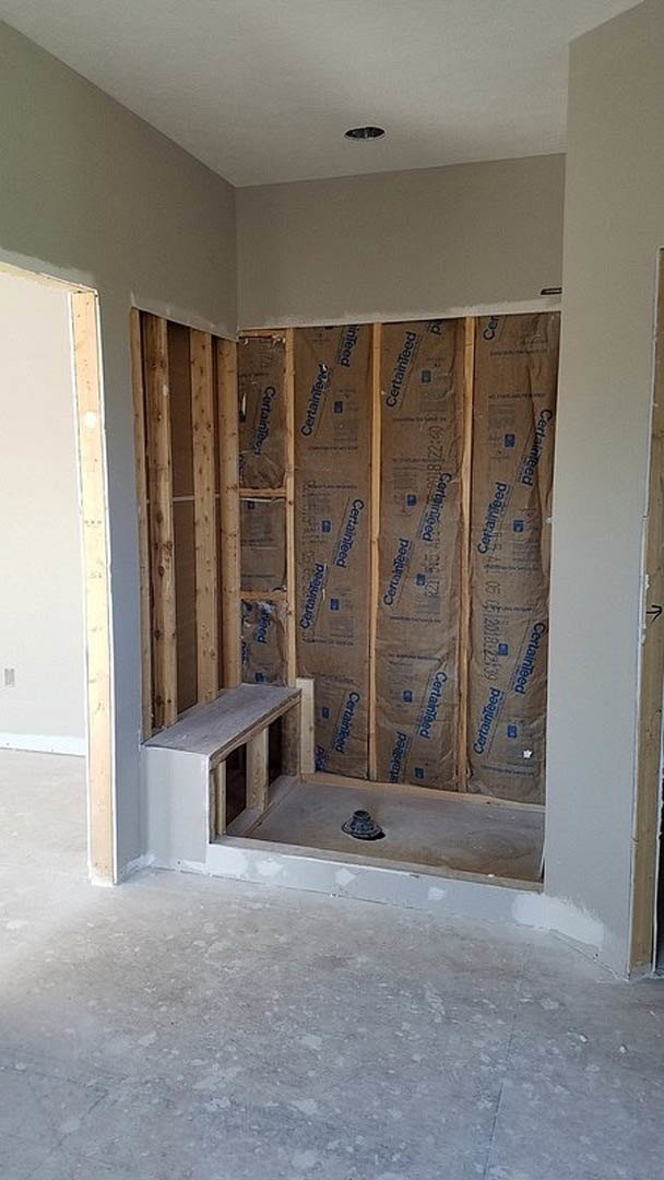 Bathroom under construction featuring exposed insulation, unfinished plaster walls with a cutout, concrete floor with central drain, and a window framed by raw materials.