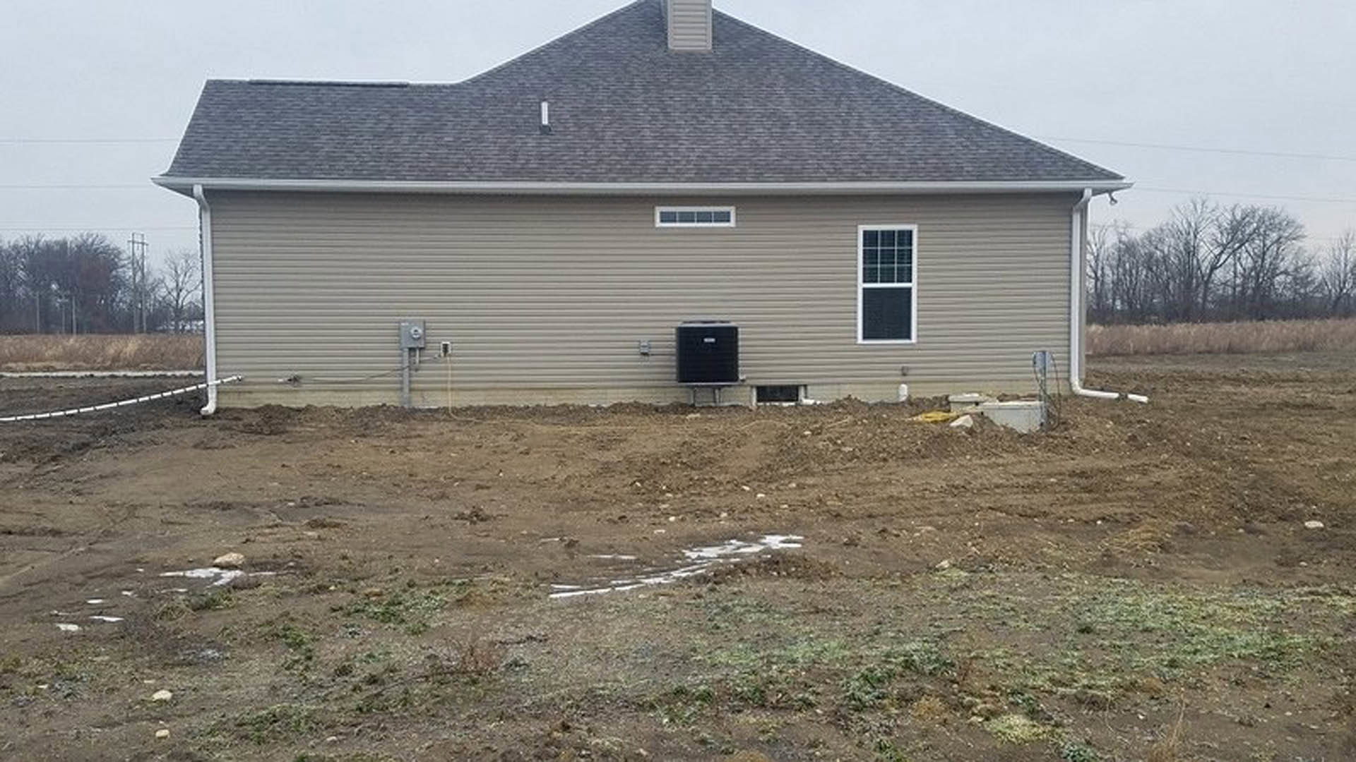 Modern cottage-style home with black siding, white-framed windows, and a muddy dirt lot in the foreground; grassy area and trees in the background.