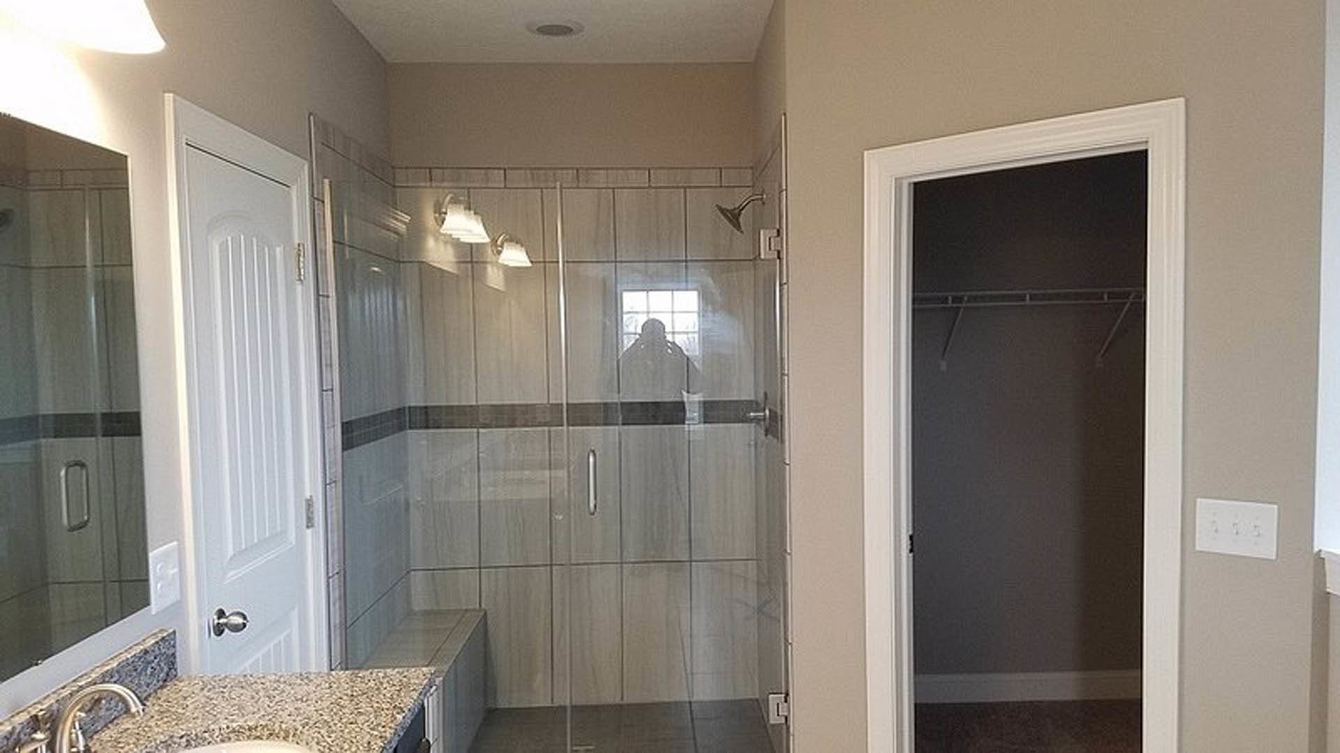 Modern bathroom featuring a frameless glass shower enclosure, large wall mirror, white ceramic sink, and gray tile walls.