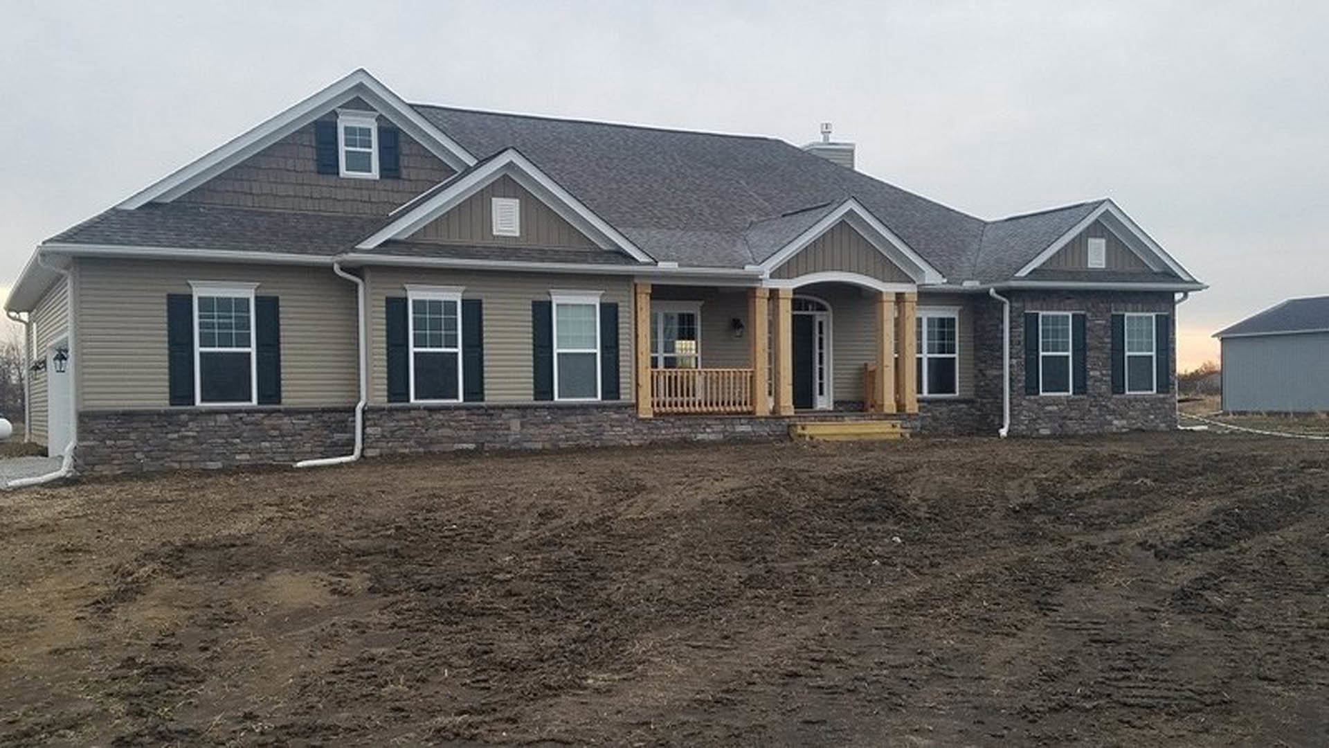 Gray-sided custom home with white trim, covered front porch, white-framed windows, and unfinished dirt yard bordered by a brick retaining wall