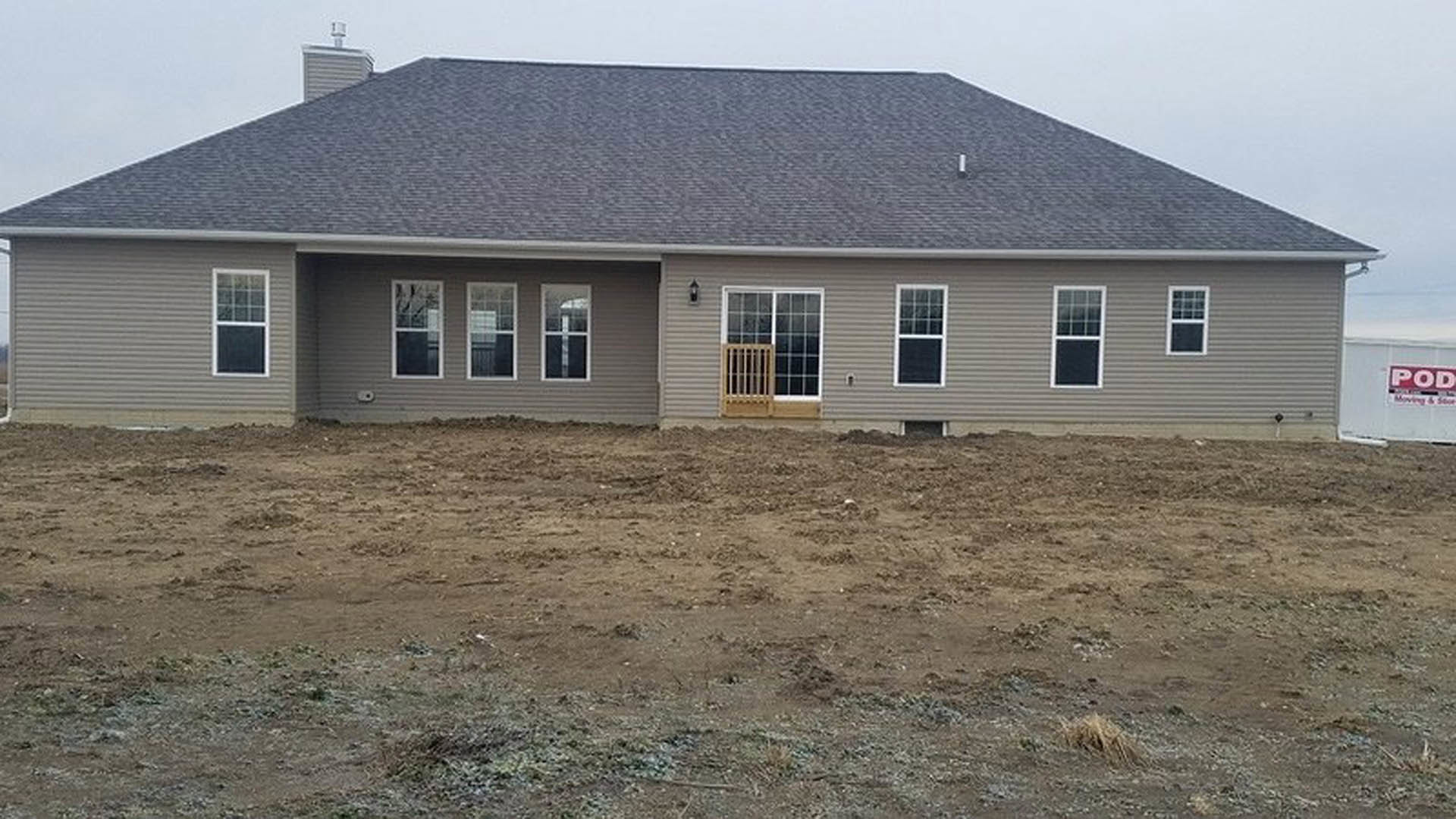 Single-story house with light siding, covered porch, white-framed windows, wooden railing, red and white sign, and dirt yard in front