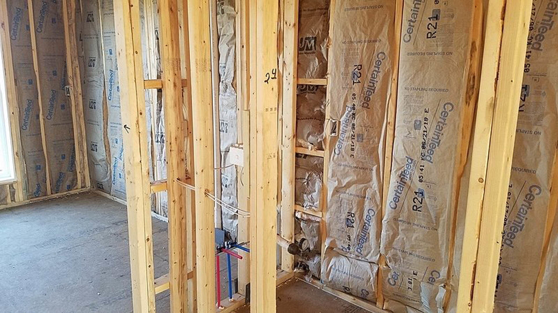 Exposed wooden framing and beams inside a partially constructed house, with insulation visible between studs and a numbered white tag attached to a wall.