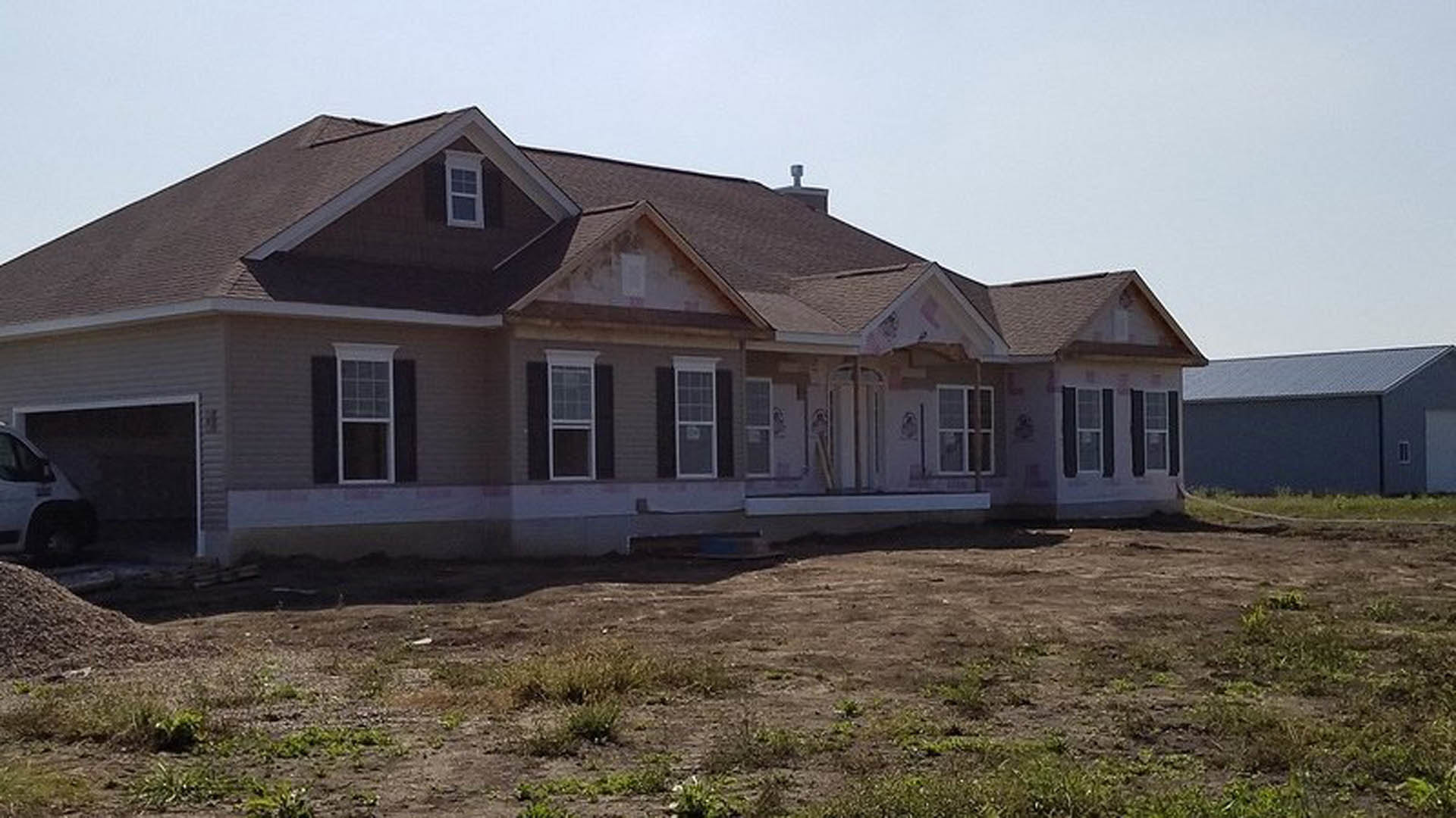 Partially built house with white-framed windows, exposed siding, and construction materials, set in a grassy dirt field with a car and another building in the background