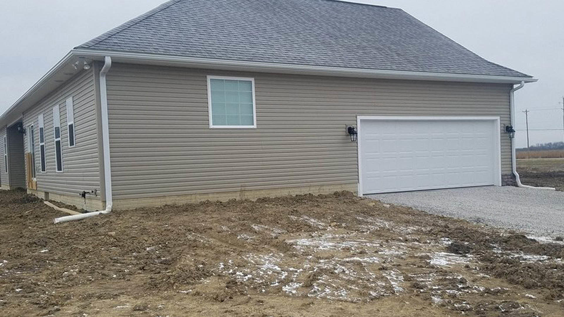 White paneled garage door with rectangular windows, white trim, gray siding, pitched roof, and patchy snow on dirt driveway.