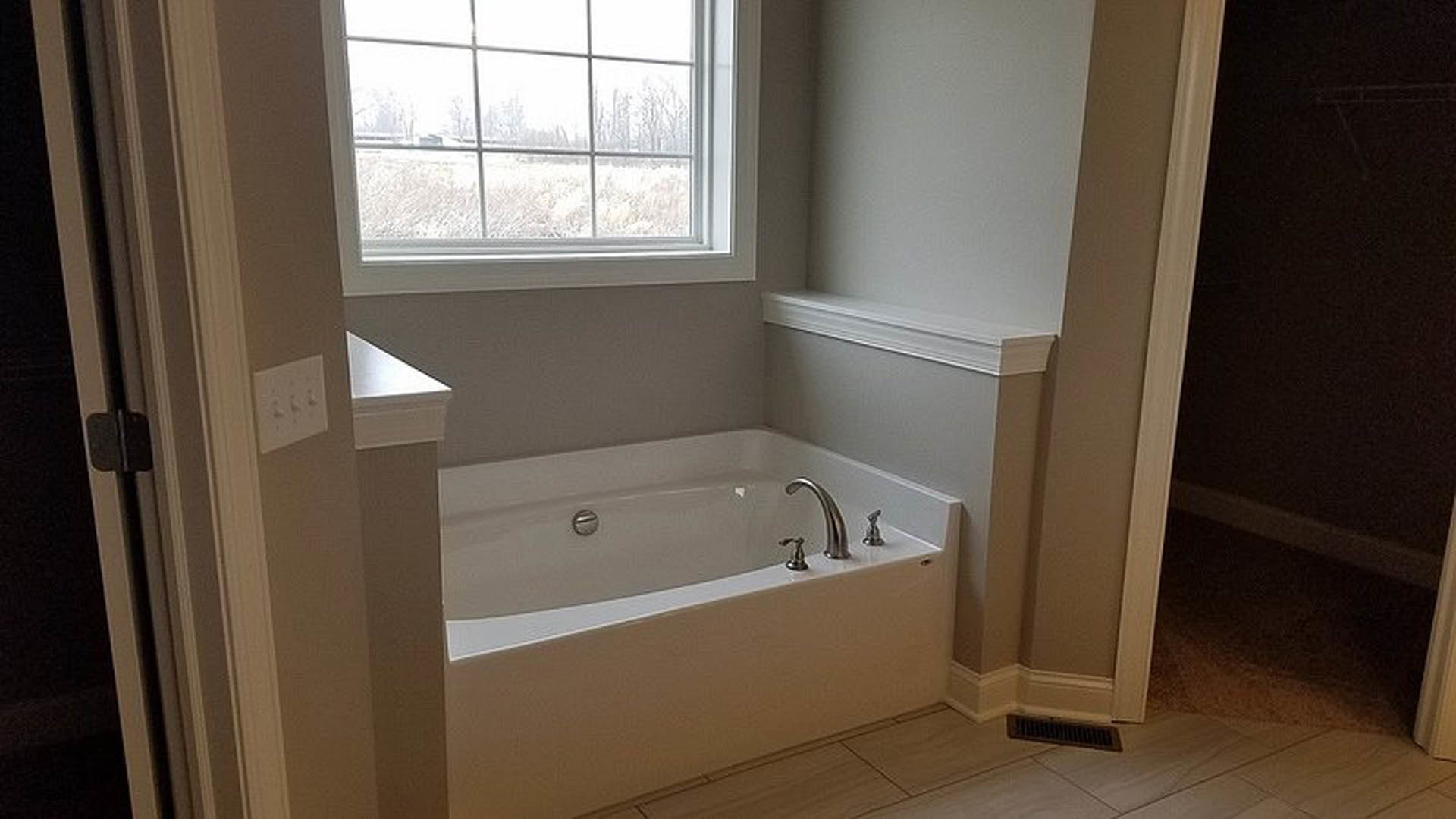 Freestanding white bathtub with chrome faucet beside large window, surrounded by light gray tile walls and floor, modern bathroom accessories visible