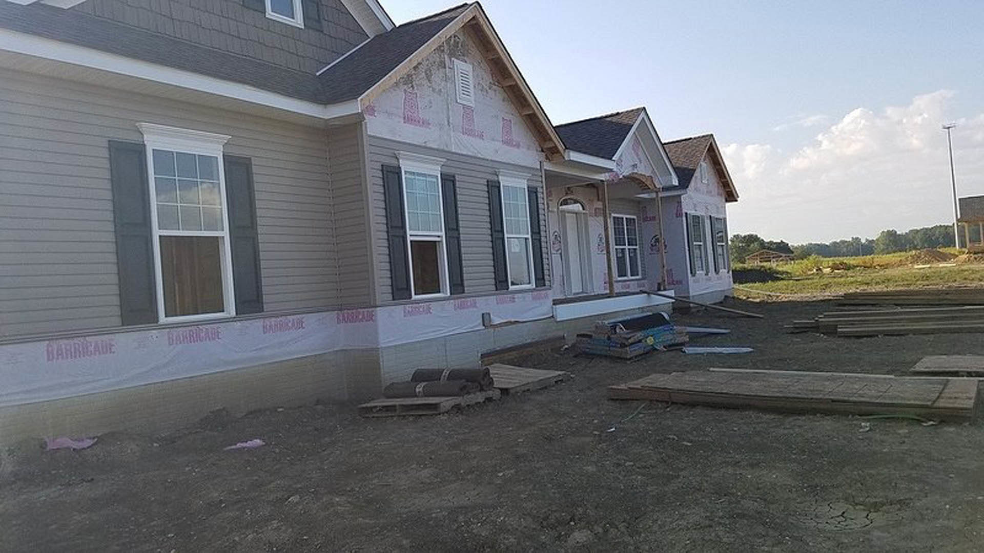 Partially built home with exposed framing, white window frames, scattered pallets on dirt ground, cloudy sky above, and light-colored siding