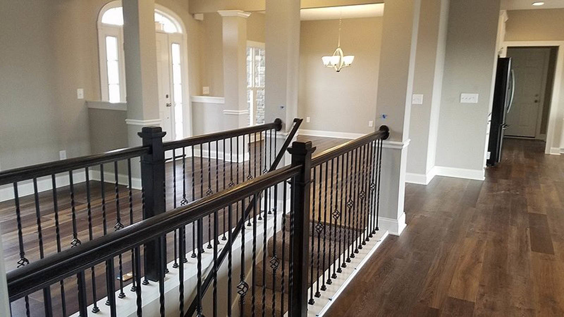 Wooden staircase with white balusters and handrail, light-colored flooring, neutral walls, and ceiling fixtures visible in a modern interior; blurred hand in foreground.