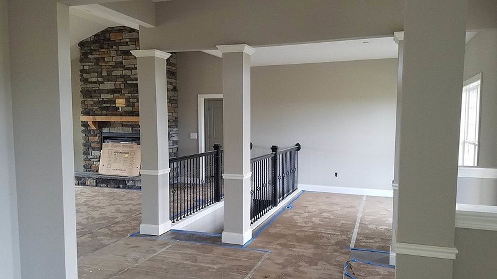 Open living room with stone fireplace, white pillar, black metal staircase railing, light wood floors, and neutral plaster walls