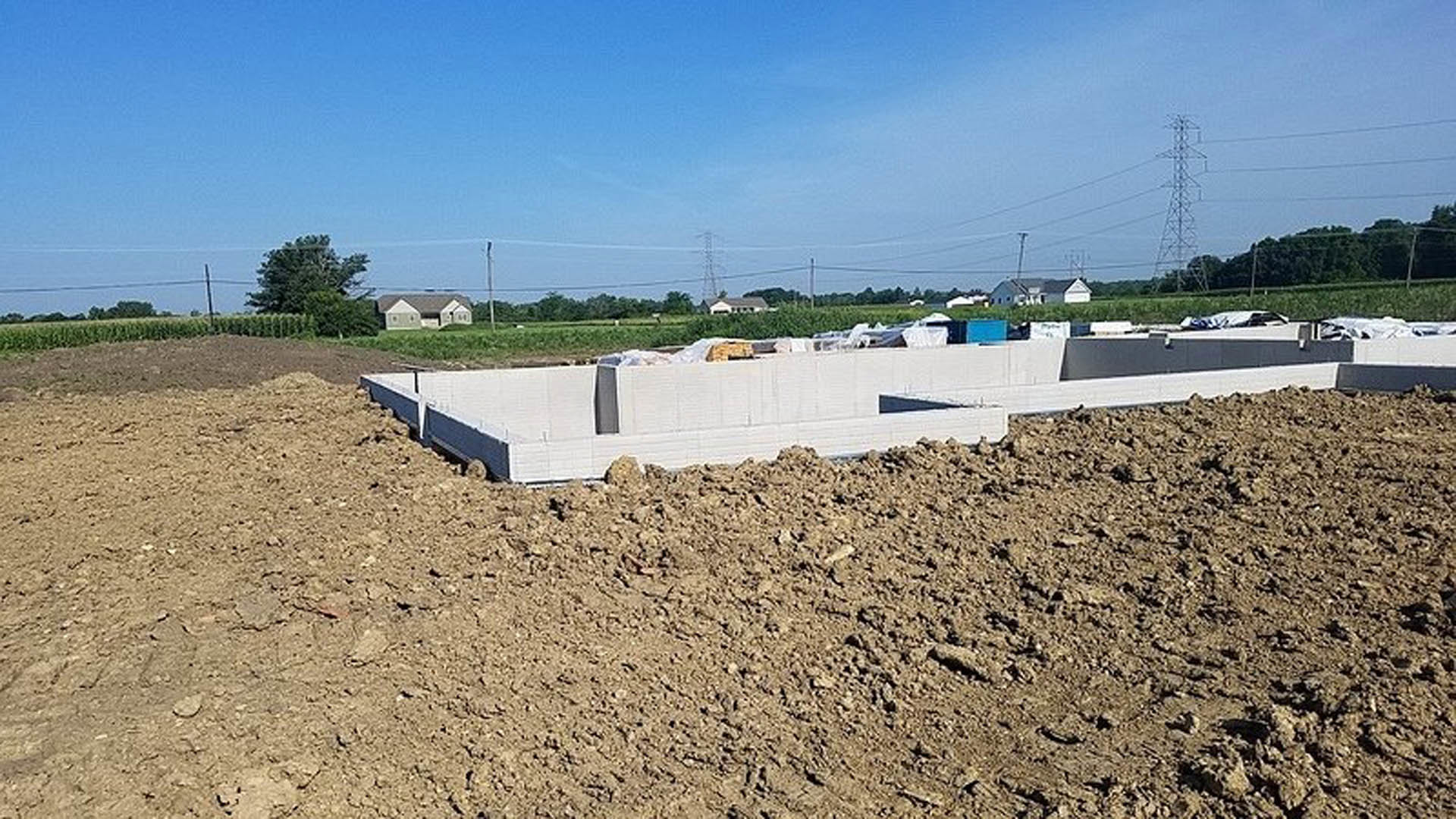 Concrete house foundation set on soil with a pile of rocks nearby, surrounded by trees and open sky
