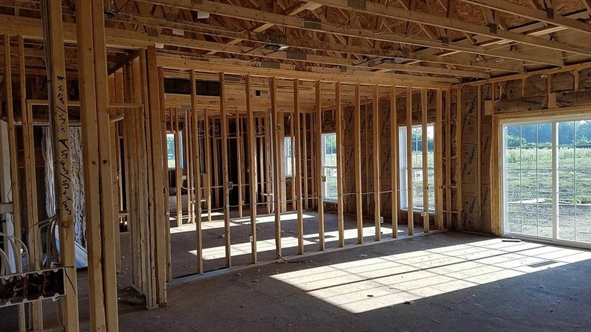 Framed room with exposed wooden beams, large windows overlooking a grassy field, unfinished plank flooring, and natural daylight streaming inside