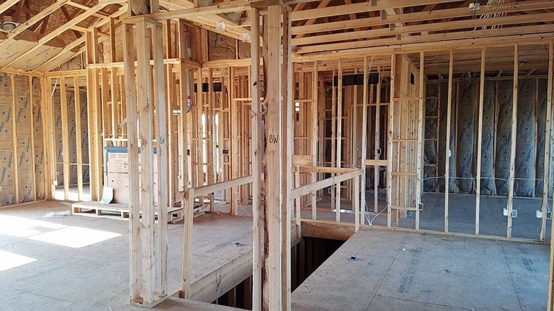 Exposed wood framing and beams with wall insulation bags, concrete floor, white tile, and partial view of a person’s head in a home under construction