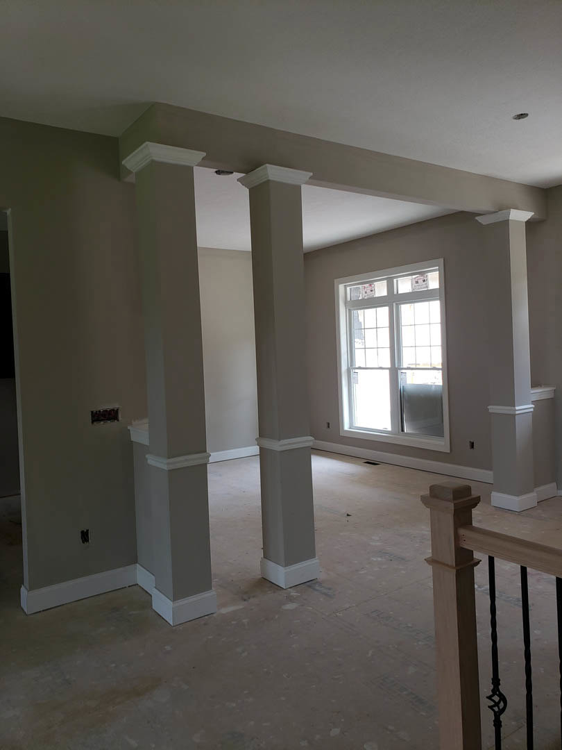 Open living area featuring white plaster columns, multi-pane window, wooden staircase with close-up railing, white walls, and white door.