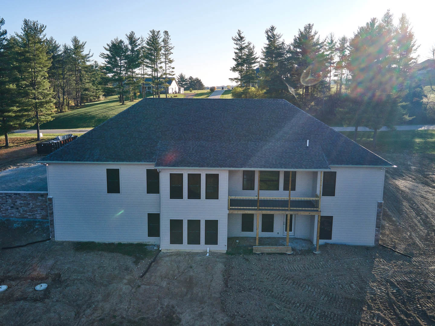 White two-story house under construction with front balcony and metal railing, surrounded by trees; row of trash cans near the entrance, unfinished roof visible.