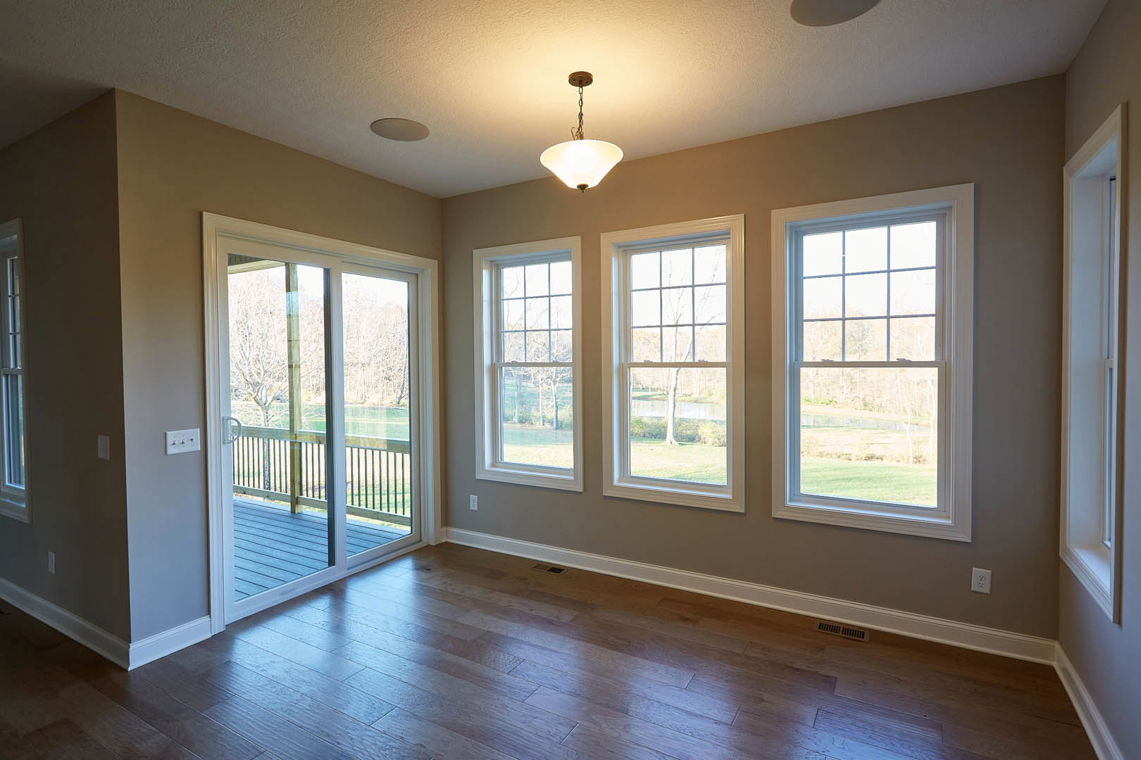 Open living area with large windows overlooking trees and a field, glass door leading to deck, hardwood floors with floor vent, ceiling-mounted light fixture